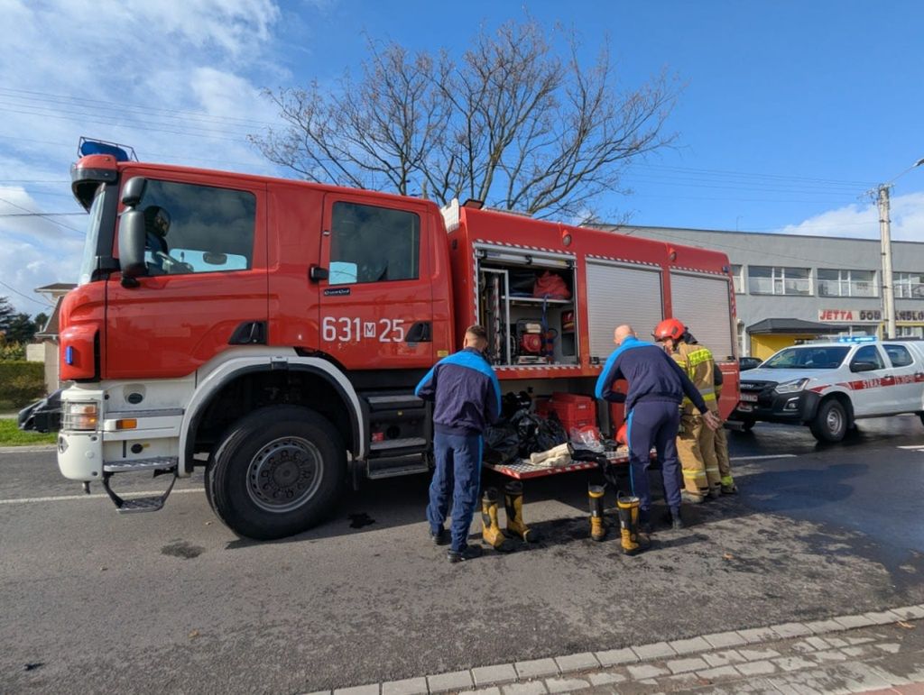 Czterech strażaków w mundurach przygotowuje sprzęt na tylnym zjeździe wozu strażackiego na ulicy w mieście.