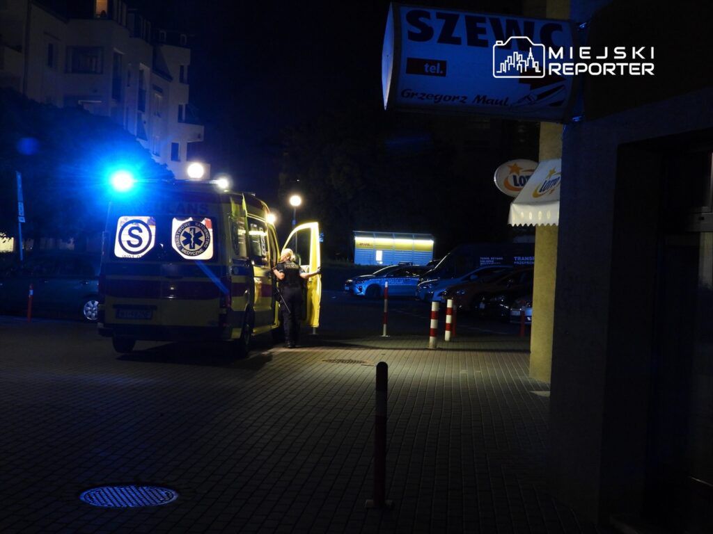 Karetka pogotowia z włączonymi sygnałami świetlnymi stoi na parkingu, a ratownik rozmawia z osobą przy otwartych drzwiach.