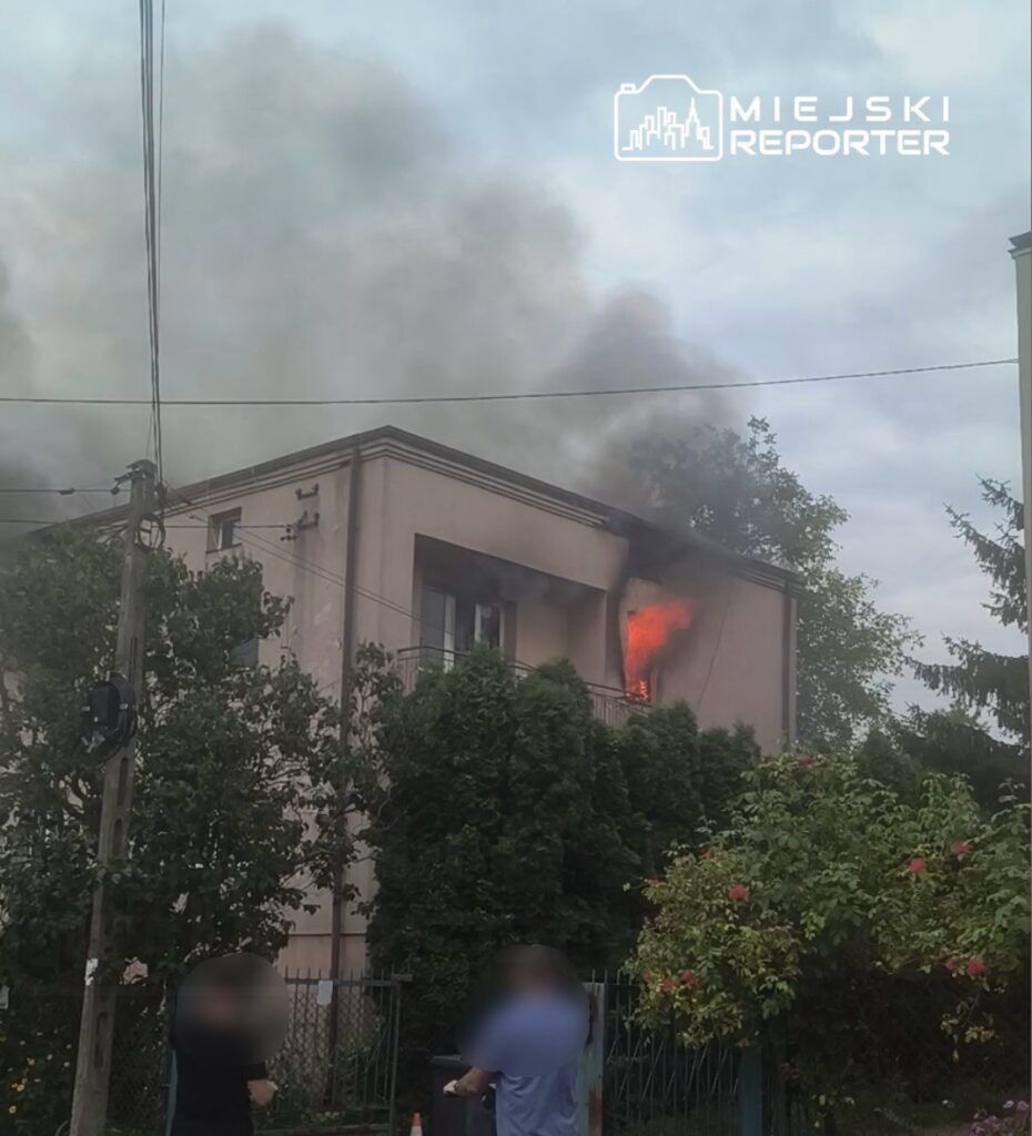 Płomienie wydobywające się z okna na piętrze budynku, w tle gęsty dym i zieleń drzew. Przy ogrodzeniu stoją dwie osoby.