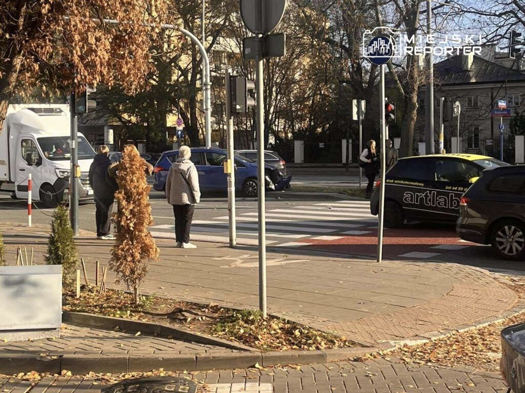 Grupa pieszych czeka na zielone światło na przejściu dla pieszych, obok ruchliwej ulicy z samochodami.