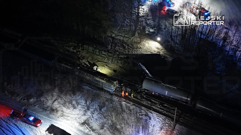 Zespół ratunkowy w odblaskowych kamizelkach bada wykolejony pociąg na zaśnieżonym torowisku w nocy.