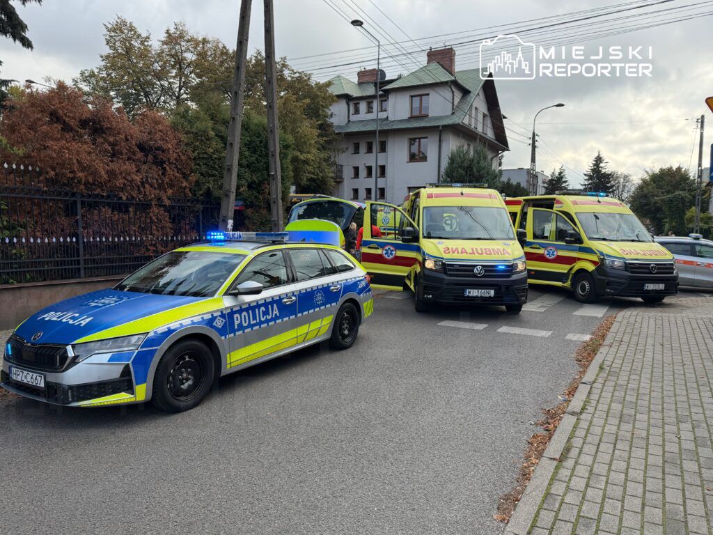 Radiowóz policji i dwa karetki pogotowia na skrzyżowaniu w miejskim otoczeniu, z włączonymi sygnałami świetlnymi.