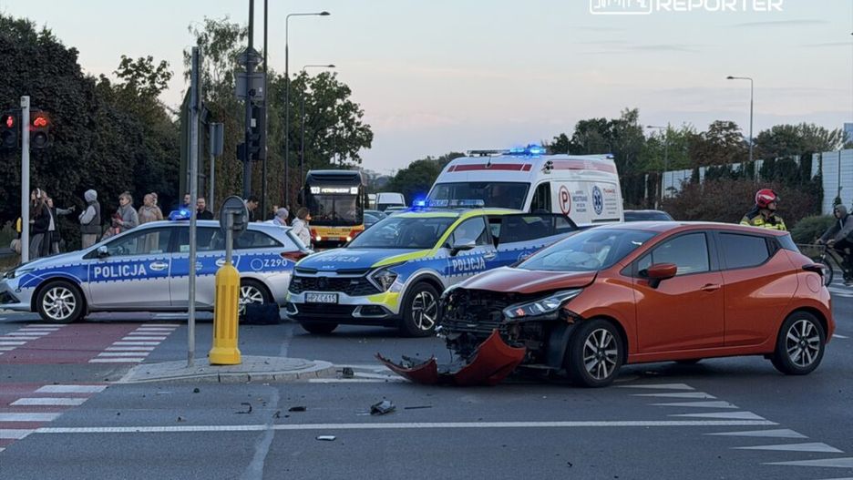 Na skrzyżowaniu stoją dwa radiowozy policji, a obok widać uszkodzony pomarańczowy samochód osobowy. W tle zespół ratunkowy i gapie obserwujący zdarzenie.