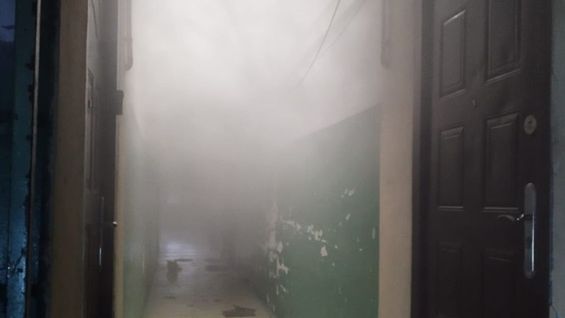 Korytarz w budynku mieszkalnym wypełniony gęstym dymem, z widocznymi drzwiami po obu stronach.