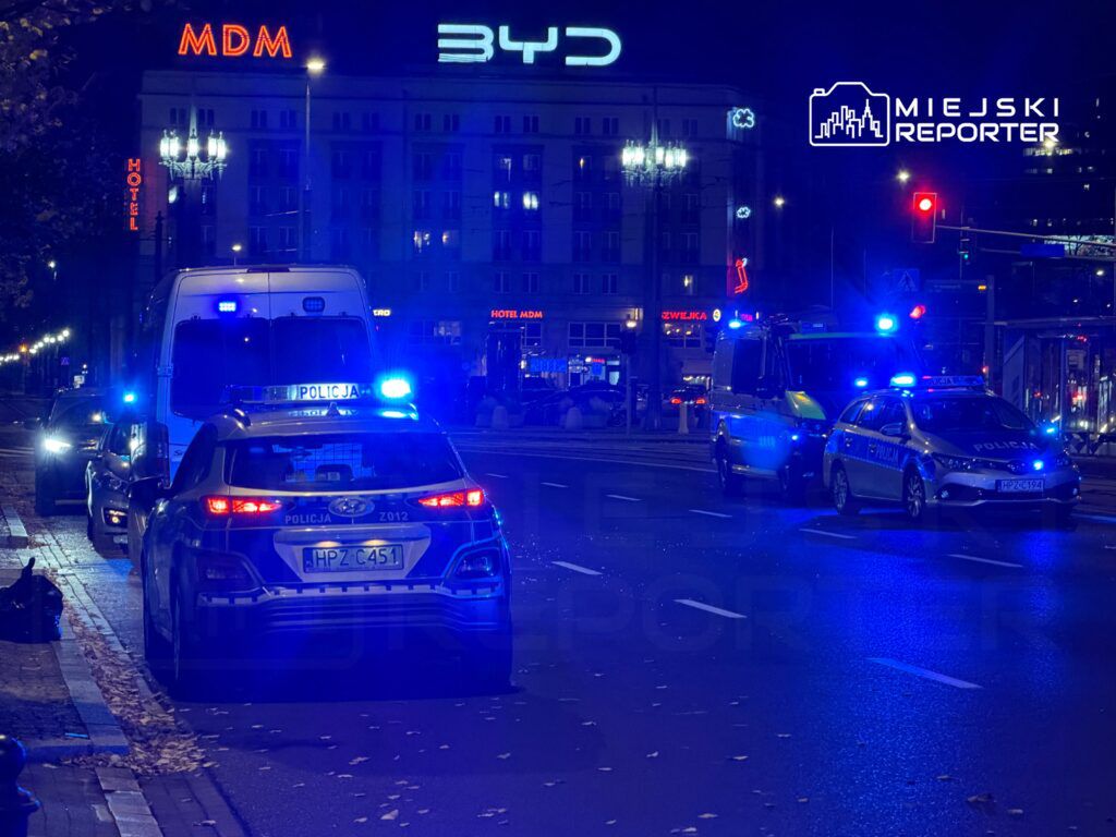 Policyjny radiowóz z włączonymi sygnałami świetlnymi stoi na ulicy nocą, obok wozu strażackiego i pojazdów cywilnych. W tle widoczny hotel z neonowym napisem.