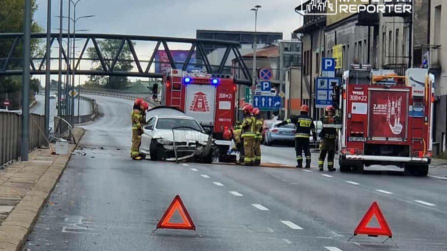 Grupa strażaków pracuje przy uszkodzonym samochodzie na jezdni, obok stoją dwa wozu strażackie.
