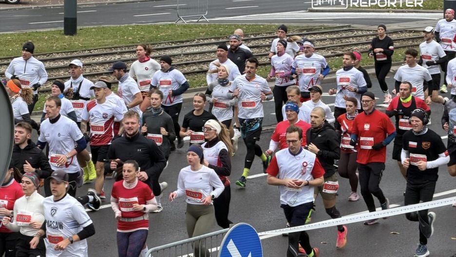 Biegacze w różnorodnych strojach biegowych uczestniczą w wyścigu na miejskiej ulicy, mijając znaki drogowe.