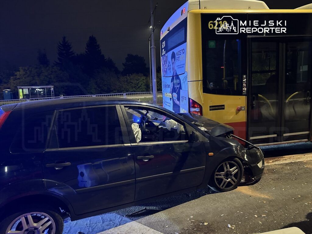 Czarny samochód osobowy z uszkodzeniami stoi obok przystanku autobusowego, obok żółtego autobusu.