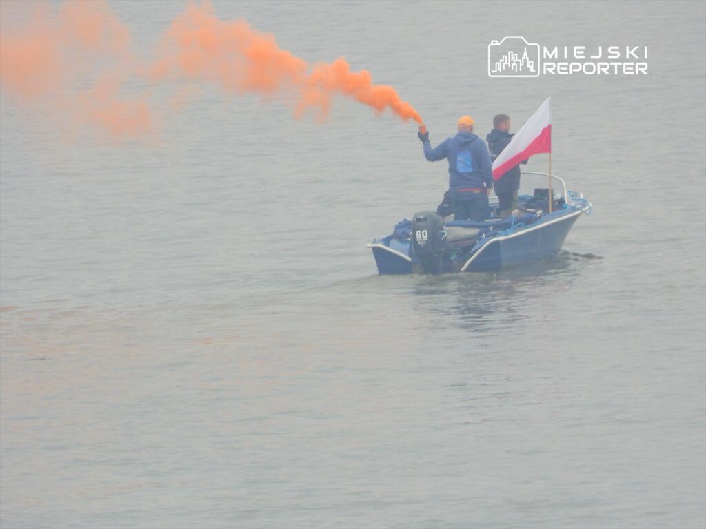 Mężczyzna w niebieskiej kurtce trzyma pomarańczowy dymny flar, a drugi mężczyzna stoi obok z flagą Polski na łodzi na wodzie.