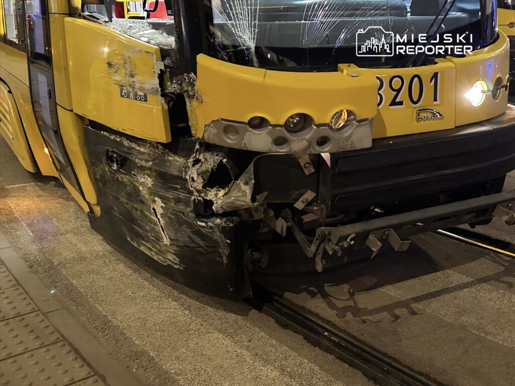 Uszkodzony tramwaj z żółtą powłoką, z widocznymi wgnieceniami i pękniętą szybą, stoi na oświetlonym torowisku.