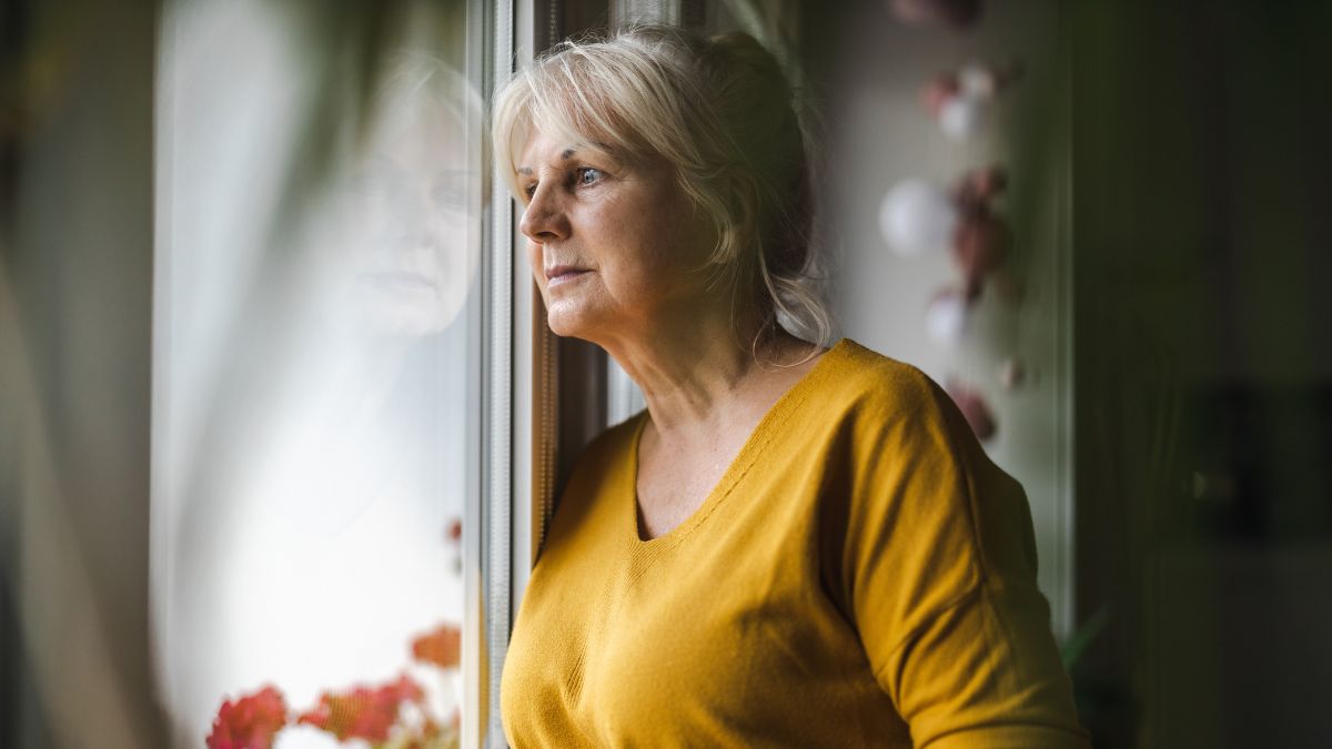 Dojrzała, smutna kobieta patrzy przez okno.