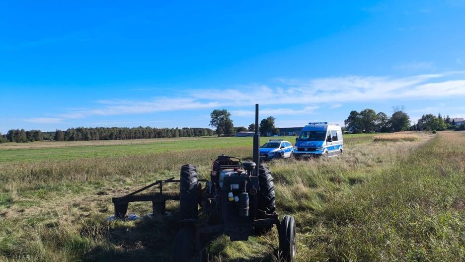 Traktor stojący na łące obok radiowozu i karetki, w tle rozległe pola i drzewa.