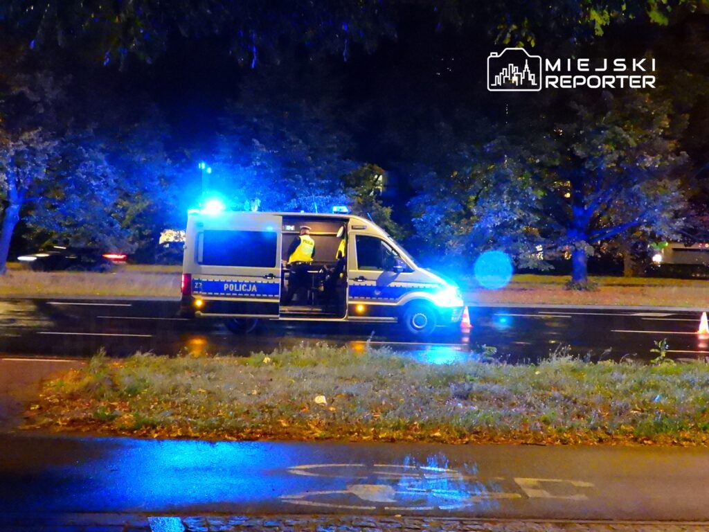 Policyjny radiowóz z włączonymi sygnałami świetlnymi stoi na poboczu drogi w nocy, obok widoczne są pachołki.