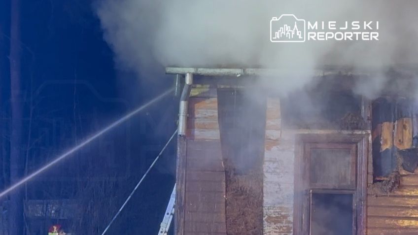 Strażak gaszący pożar na dachu drewnianego budynku, z którego wydobywa się gęsty dym.