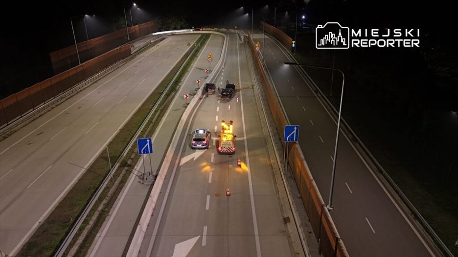 Roboty drogowe prowadzone na nocnej jezdni, z oznakowaniem i pojazdem zabezpieczającym w akcji.