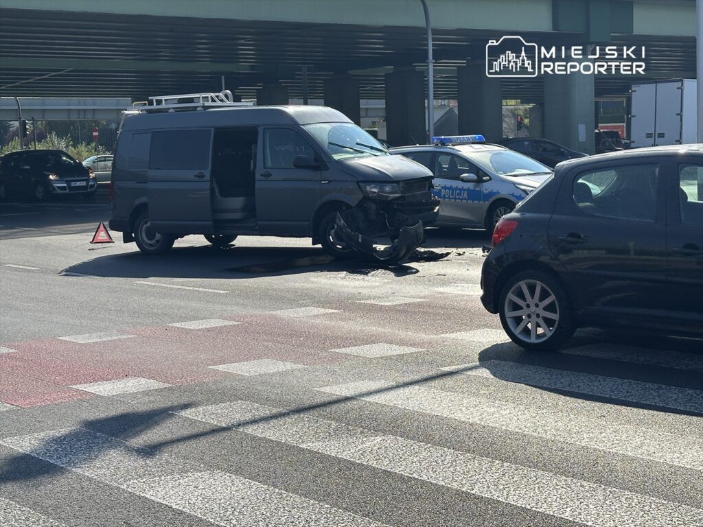 Zderzenie dwóch pojazdów na skrzyżowaniu, z uszkodzonym vanem i radiowozem policji w tle.