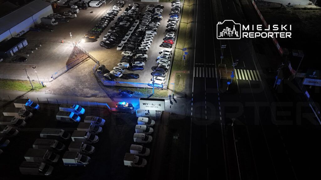 Zespół policji w niebieskich mundurach prowadzi czynności na terenie parkingu, otoczonego zaparkowanymi pojazdami.