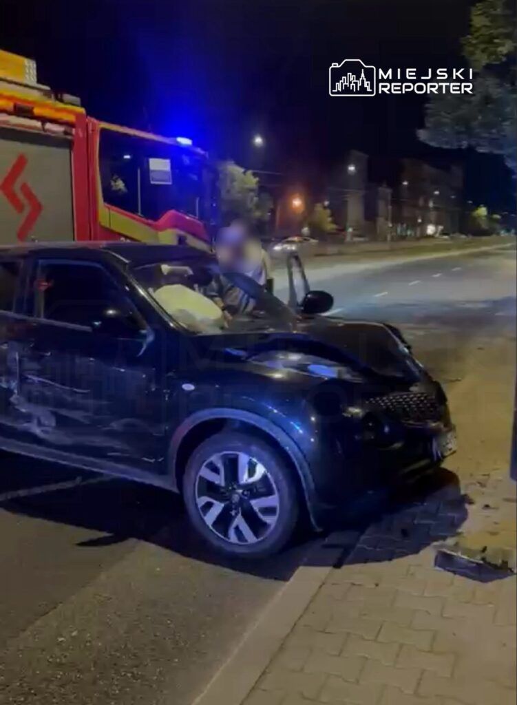 Czarny samochód osobowy z uszkodzoną przednią częścią stoi przy drodze, obok wozu strażackiego z włączonymi sygnalizacjami.