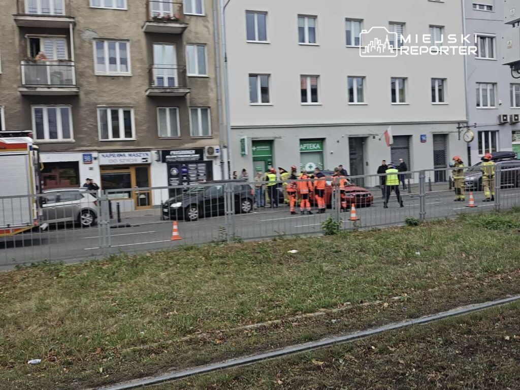 Grupa ratowników w pomarańczowych uniformach pracuje przy uszkodzonym pojeździe na ulicy, otoczona przez gapiów.