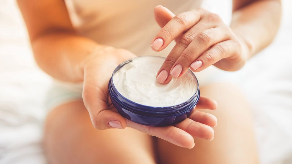 Cropped image of beautiful young woman applying cream while sitting on bed at home