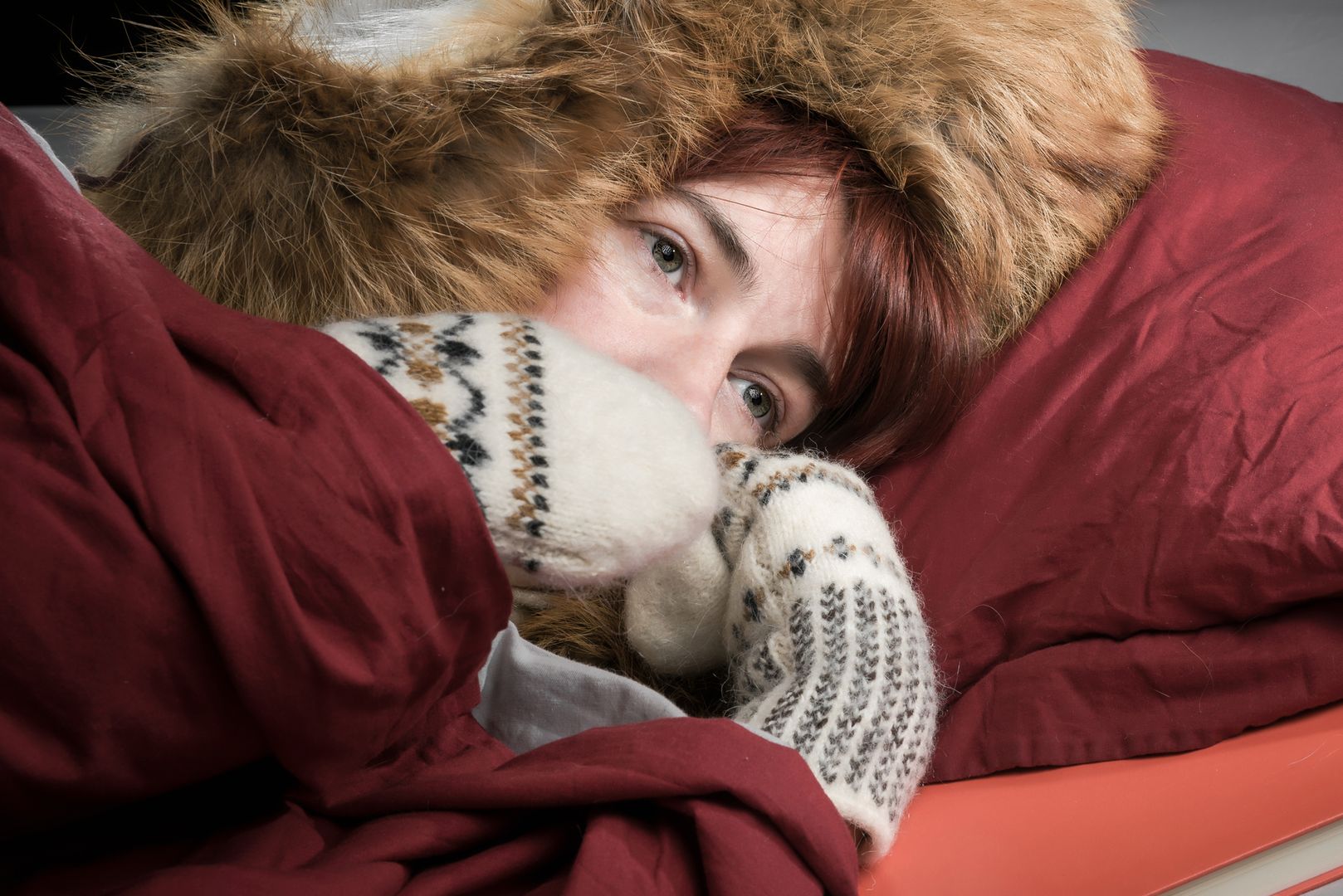 Woman lying in a bed wearing fur hat and mittens and freezing.