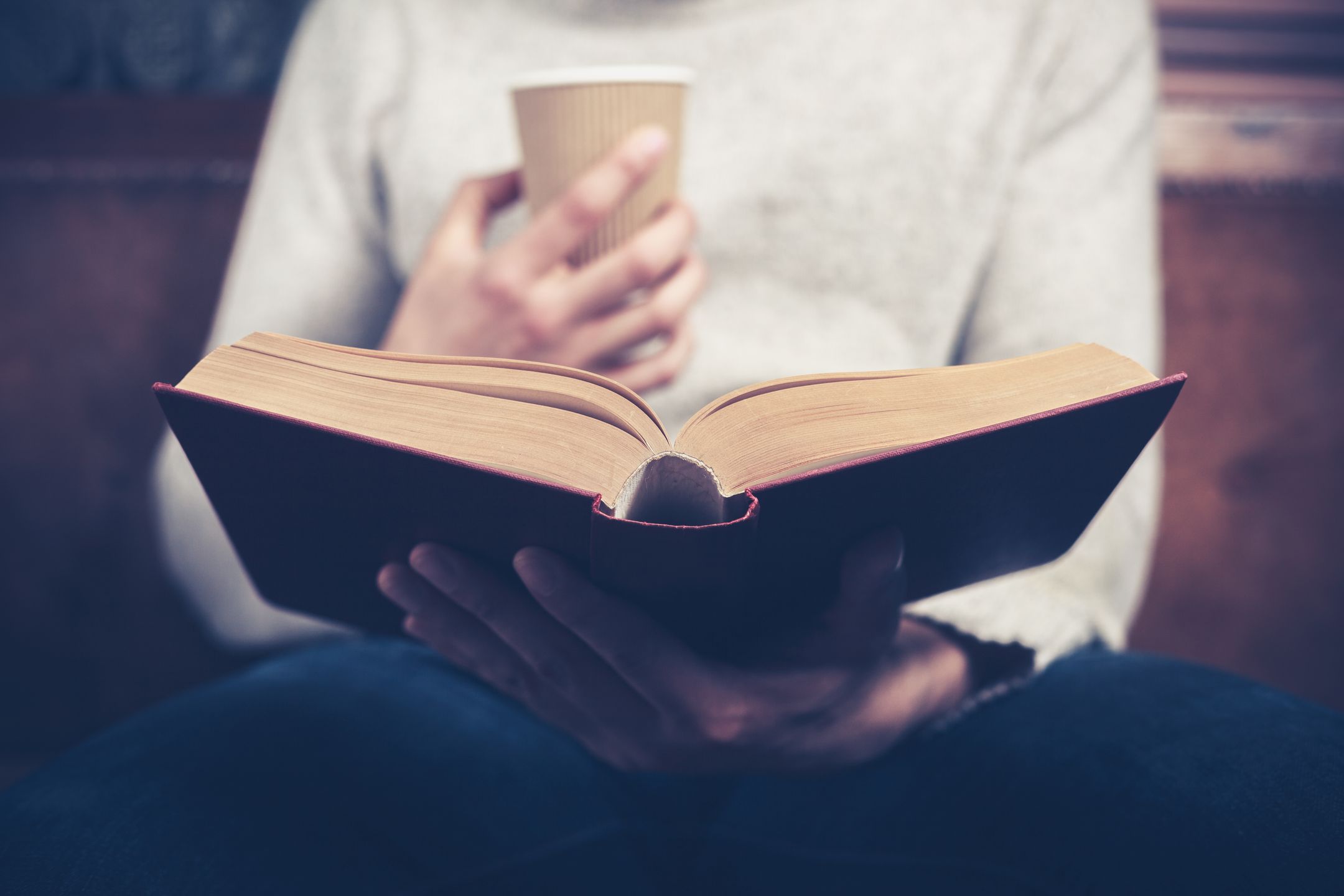 Young man is sitting on a sofa and reading a book while holding a paper cup 