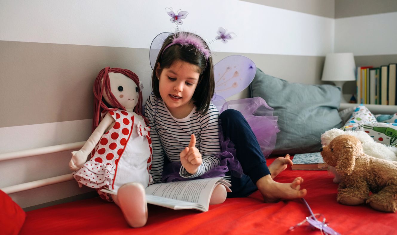 Little girl disguised as a butterfly sitting on the bed reading a book to her rag doll 