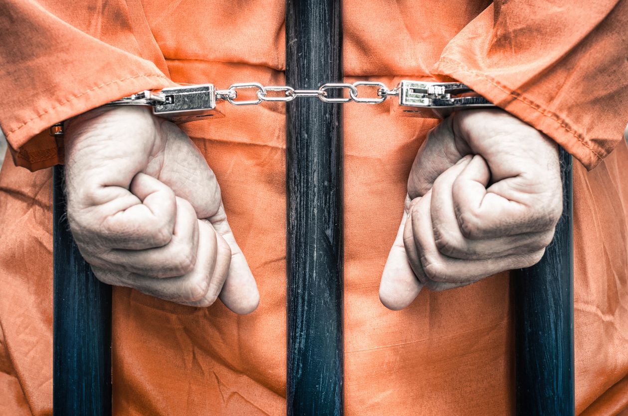Handcuffed hands of a prisoner behind the bars of a prison with orange clothes - Crispy desaturated dramatic filtered look 