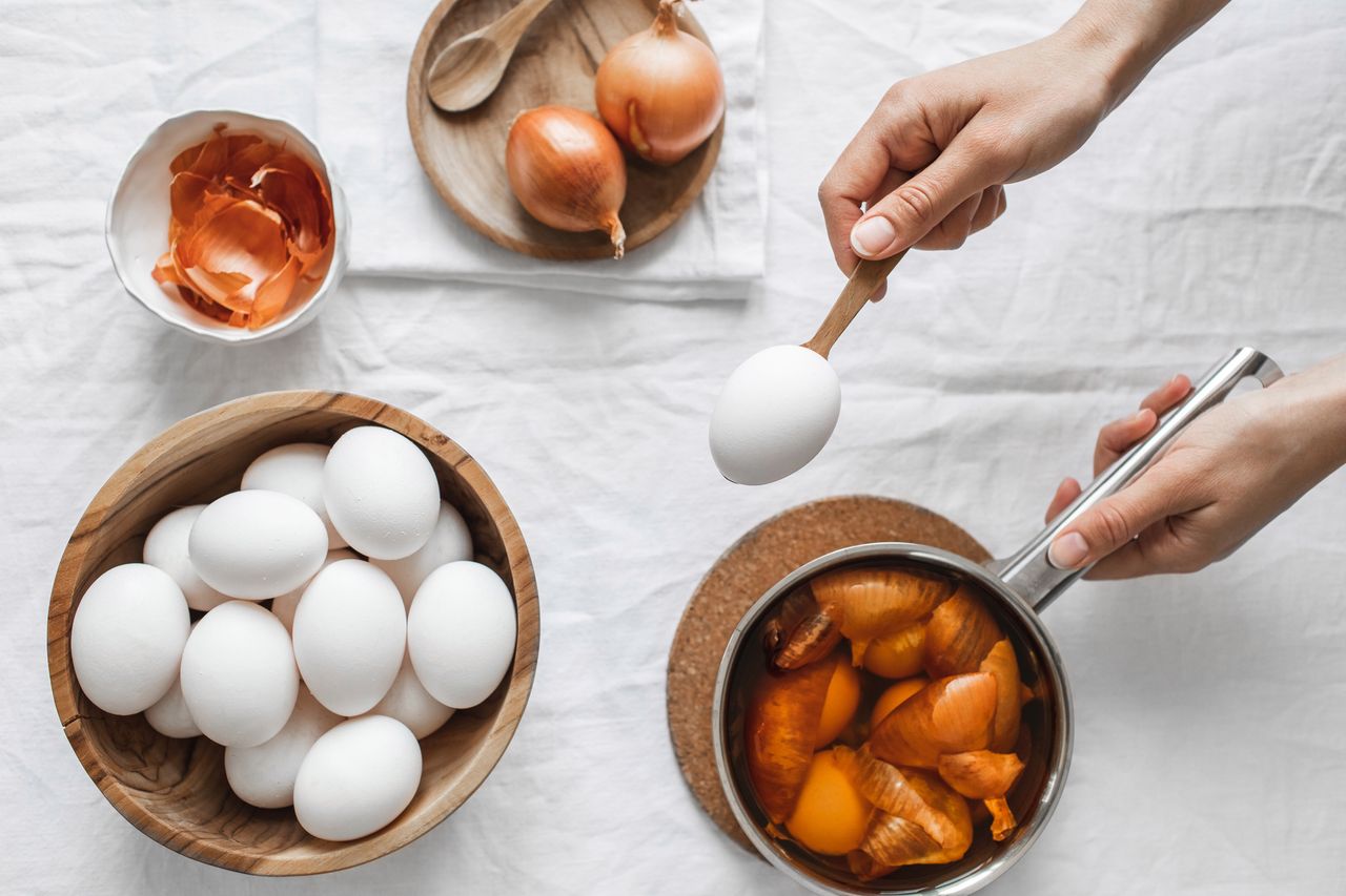Preparing for celebrating Easter. Sustainable way of painting eggs. Woman hands holding egg, Ingredients for cooking on white textile table linen background, top view. Wooden cutlery and plates. Flat lay. Zero waste concept.