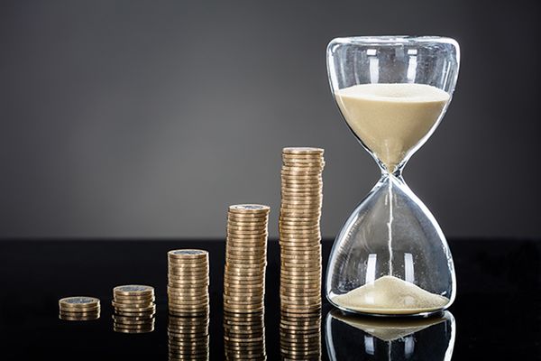 Close-up Of An Hourglass Near Stack Of Coins Over Gray Background 