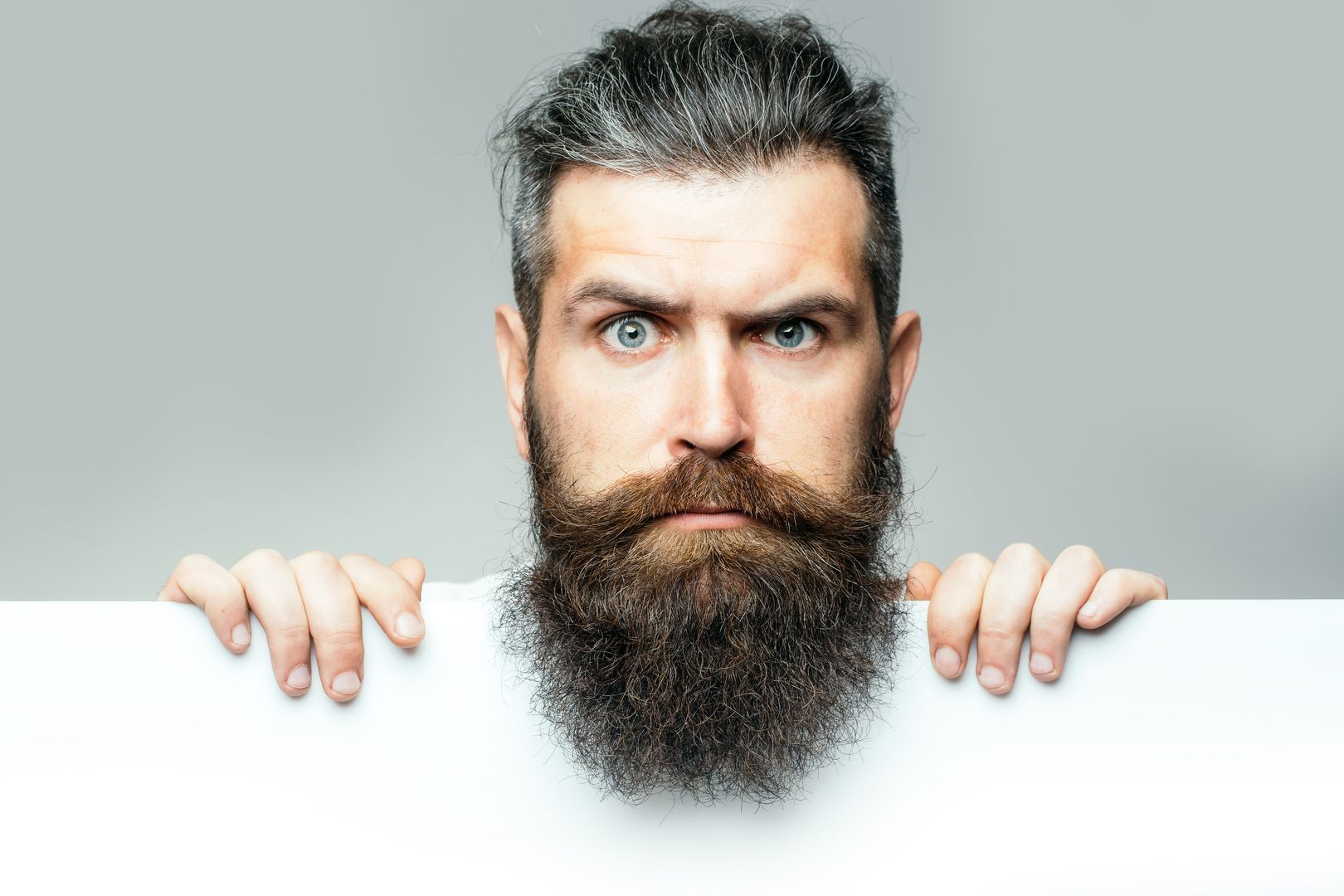 handsome bearded man with long lush beard and moustache on surprised face with white paper sheet in studio on grey background, copy space