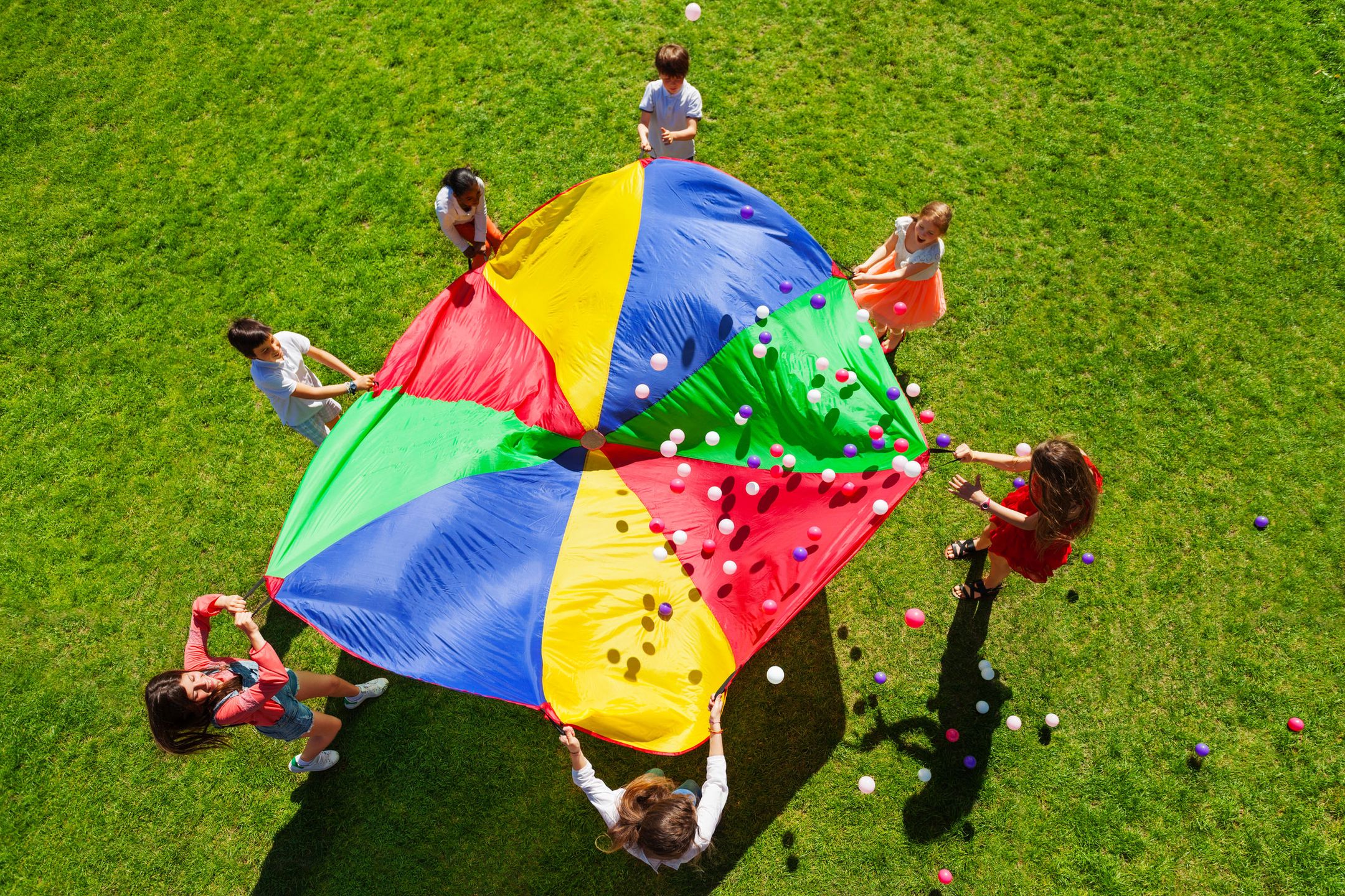 Top view picture of kids standing in a circle on the green lawn and holding rainbow parachute full of colorful balls 