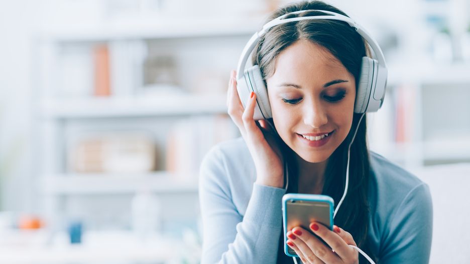 Smiling girl relaxing at home, she is playing music using a smartphone and wearing white headphones