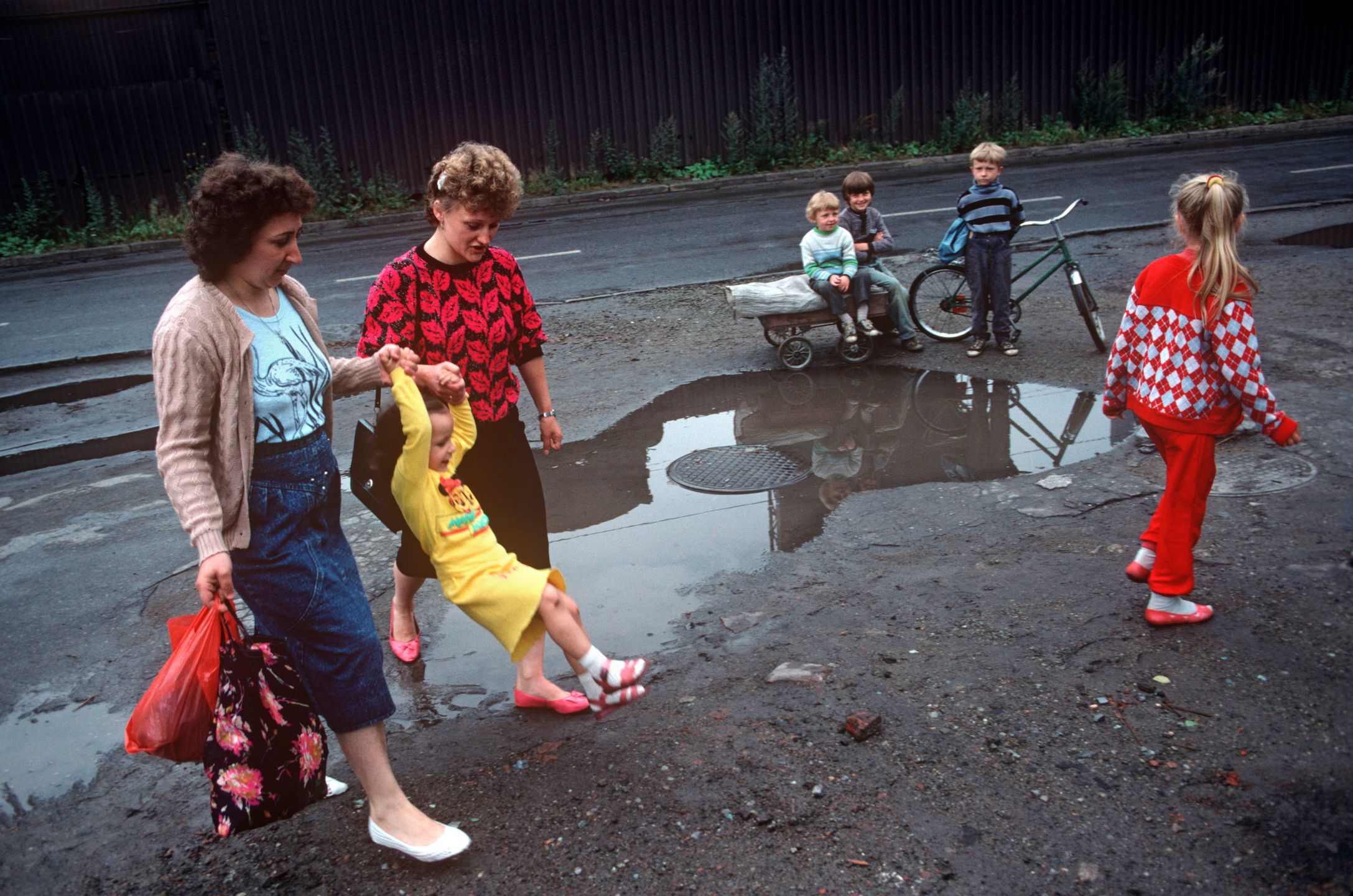Mothers swing a child across a filthy puddle in the town of Nova Huta, an industrial Stalinist steel town in southern Poland. In filthy this industrial street, three street urchins watch the colourfully-dressed girls pass-by. It is horribly depressing and unhealthy place to grow up and these children are pale and yet seem happy, with smiles on their faces. After the Second World War, Stalin decided to build an industrial Communist fantasy just outside Krakow: a model town and immense steelworks of the future. The steelworks was named after Lenin and the town named Nowa Huta - New Steel Mill. At its peak, 27,000 people worked at the Lenin Steelworks. But Solidarity grew strong forcing strikes over pay and recognition over their union. Today, it is an economic and ecological disaster area. 