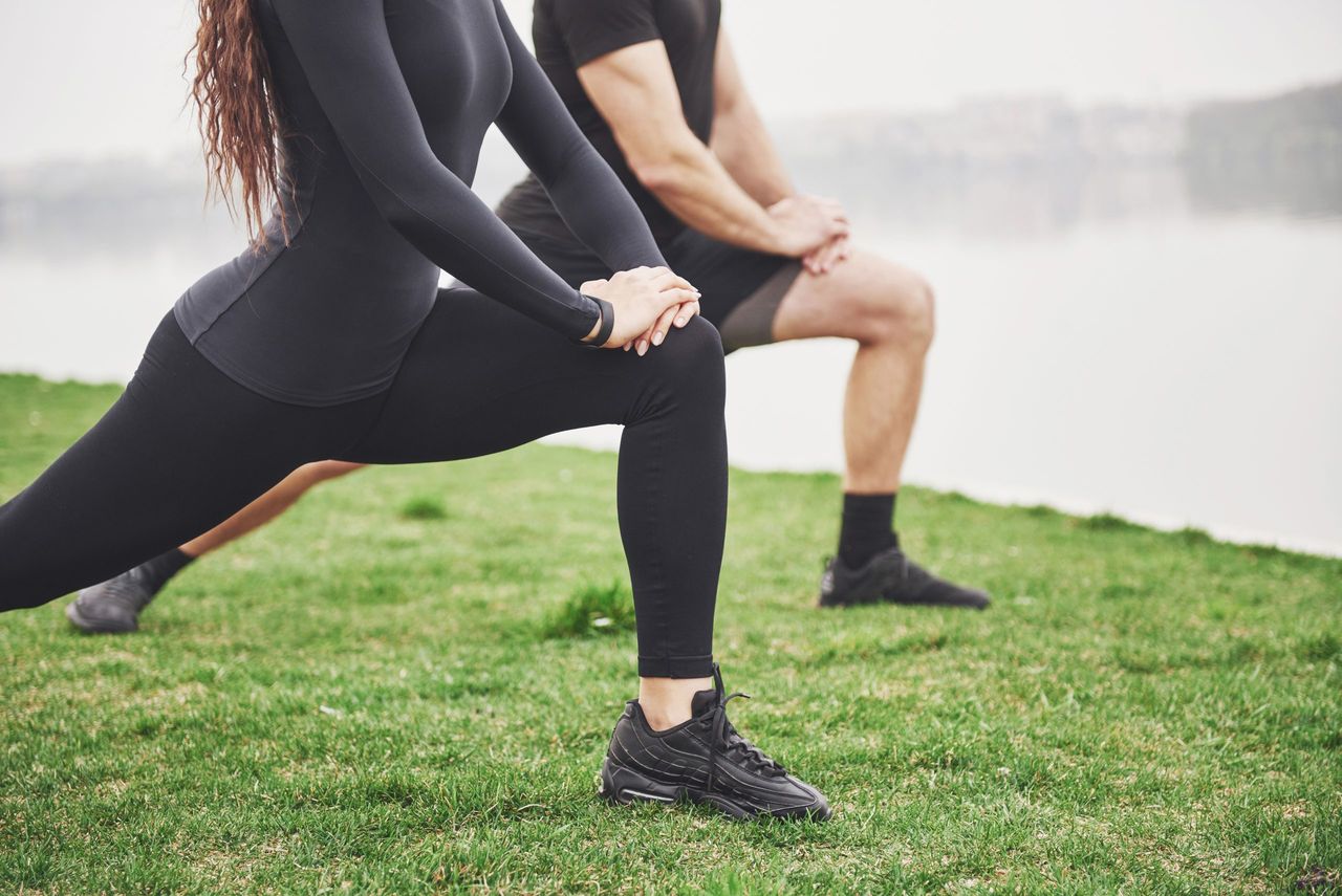 Fitness couple stretching outdoors in park near the water. Young bearded man and woman exercising together in morning.