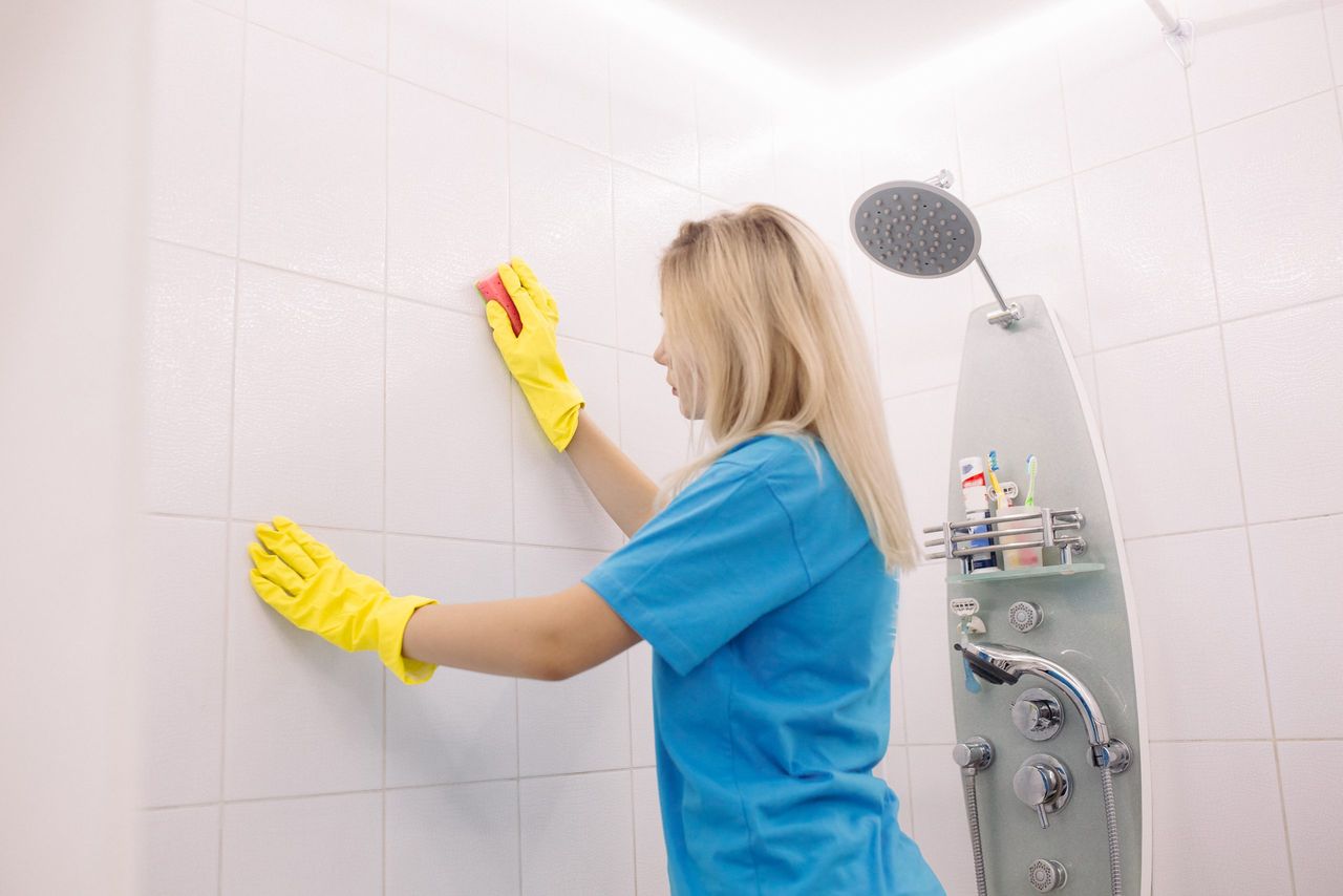 Blonde woman, employee cleaning company, wearing in yellow protective rubber gloves, with special cleaning sponge, cleaning wall tiled surface in white bathroom. Housework and housekeeping concept.