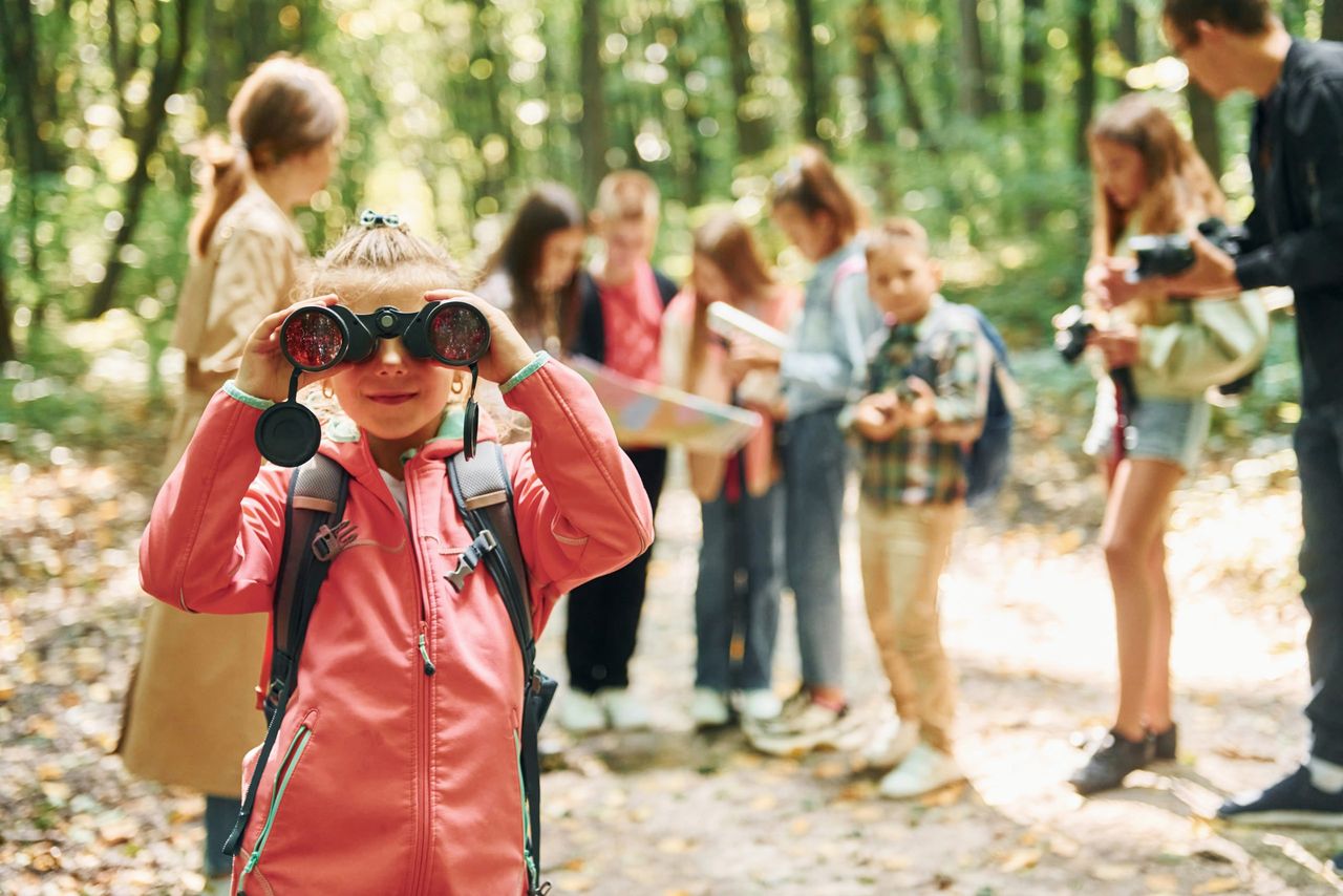 Girl looking into binoculars. Kids in green forest at summer daytime together.