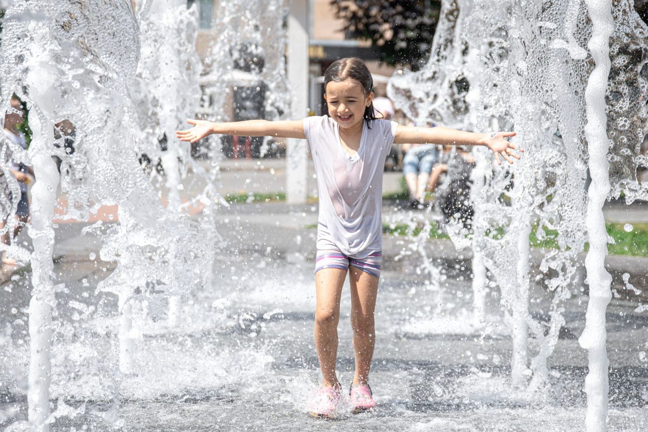 Funny little girl in a fountain, among the splashes of water on a hot summer day.