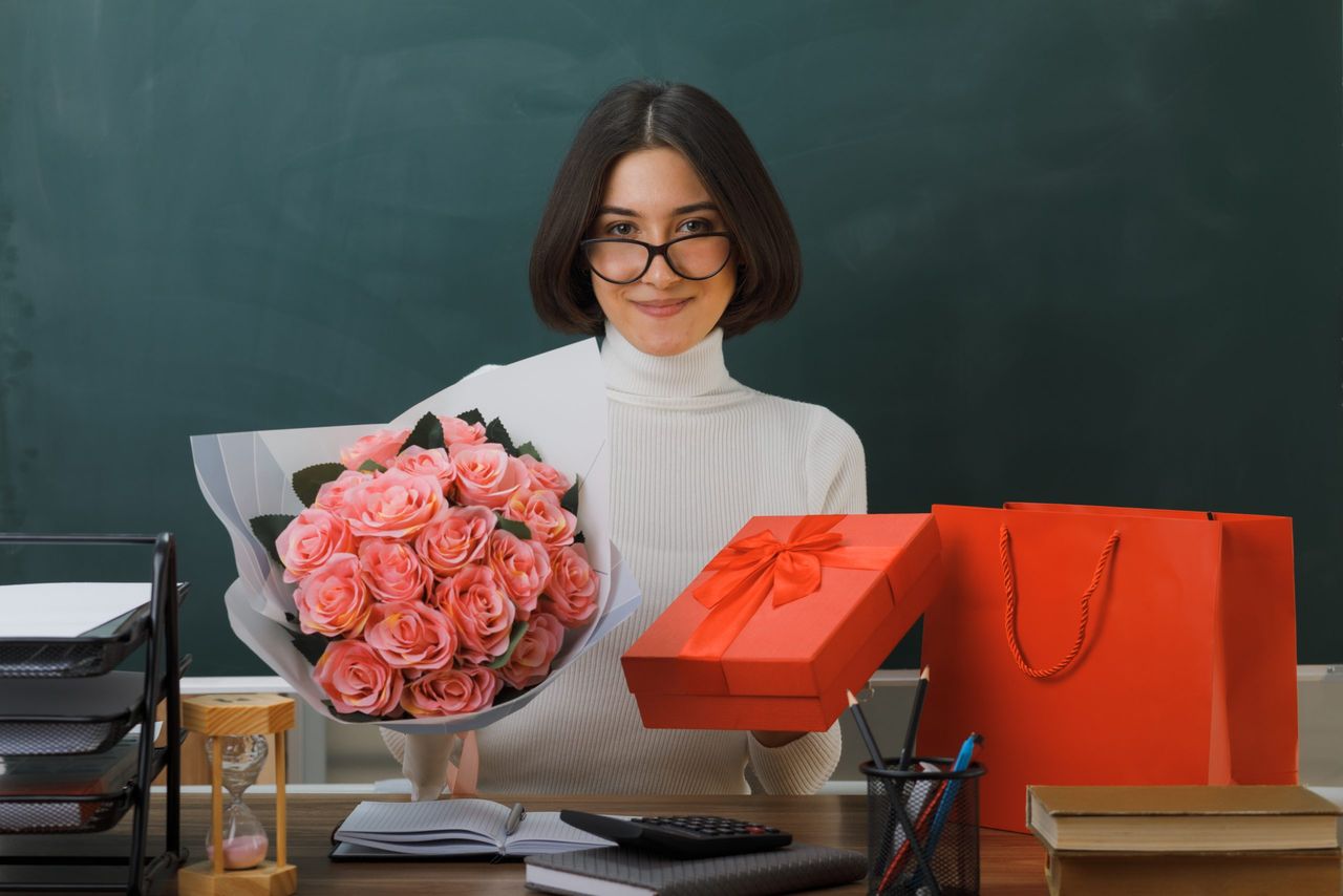 pleased young female teacher holding flowers with gift sitting at desk with school tools in classroom