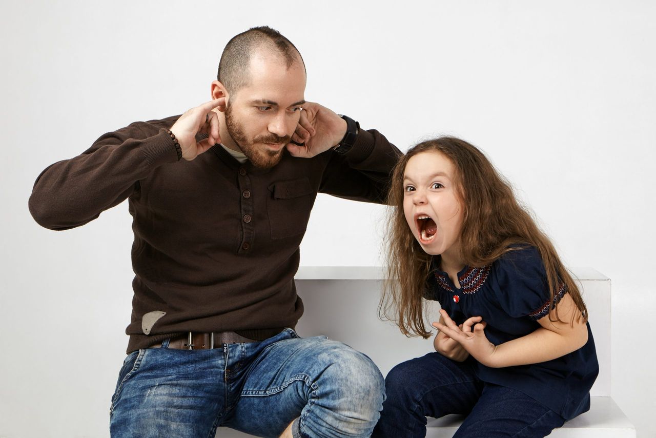 Angry little girl with long loose hair shouting, misbehaving because father didn't allow her watching cartoons. Frustrated young bearded man plugging ears, can't stand annoying screams by his daughter