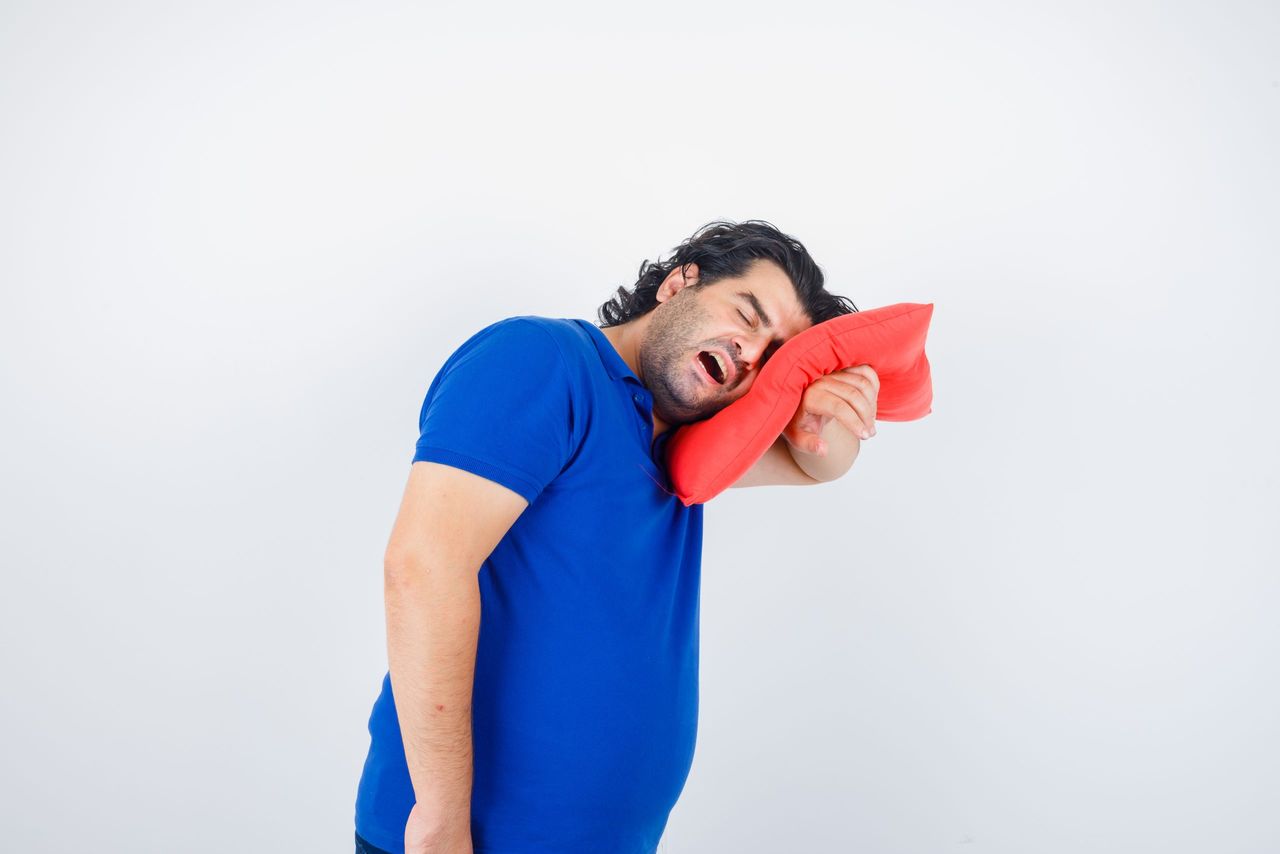 mature man in blue t-shirt leaning head on pillow while yawning and looking sleepy , front view.