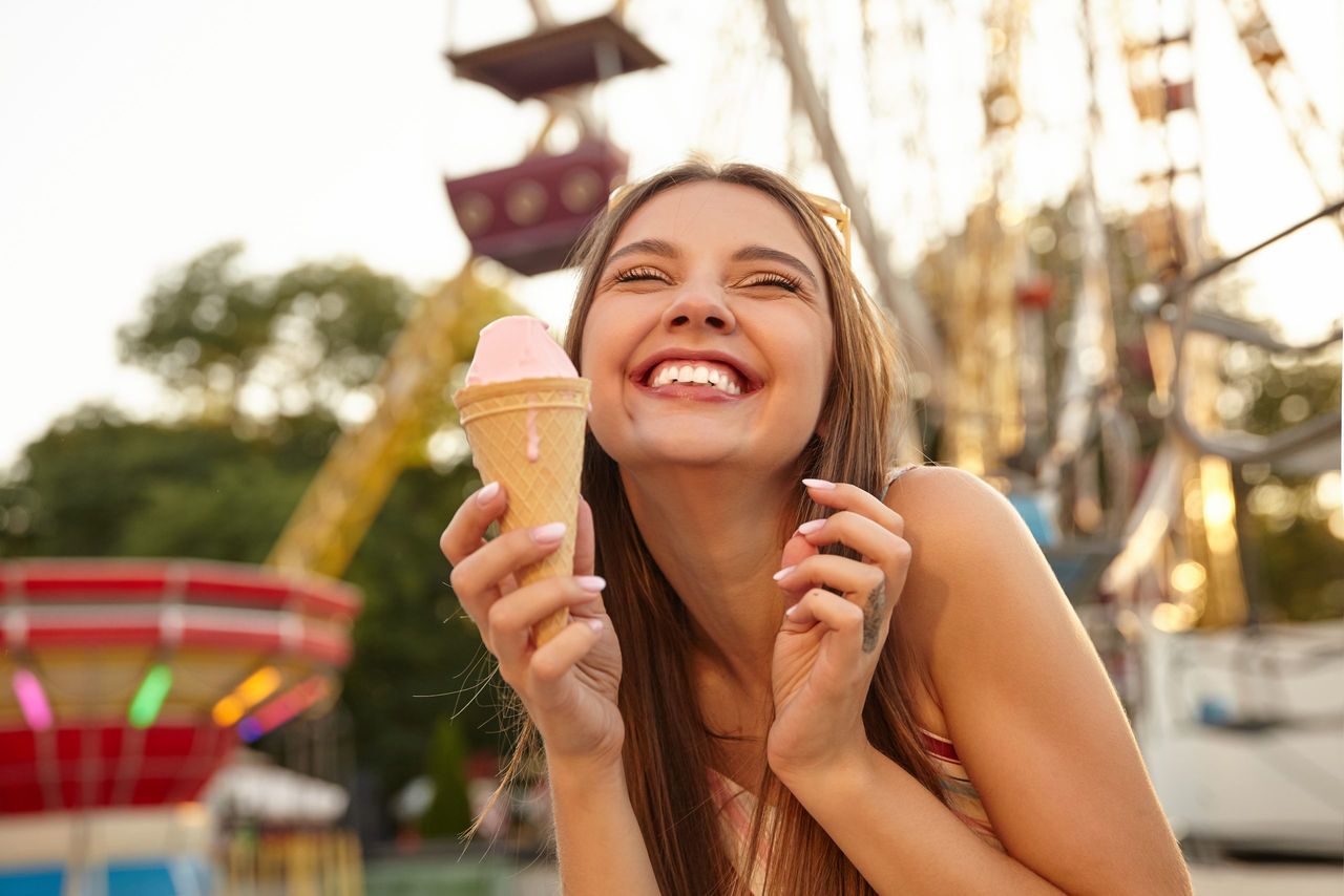 Close-up of charming cheerful young brunette lady with sunglasses on her head, showing teeth and smiling happily while eating pink ice cream cone over amusement park