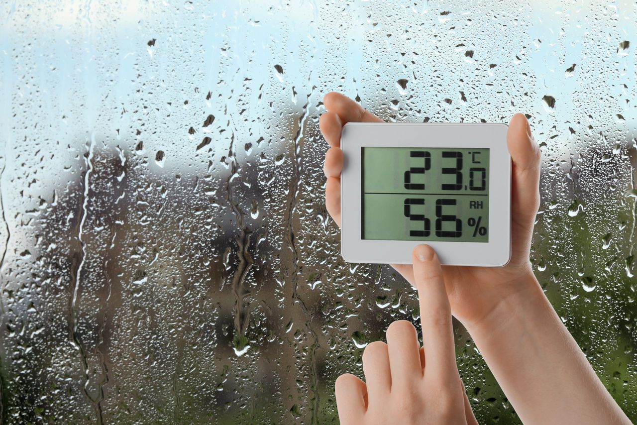 Woman holding digital hygrometer with thermometer near window on rainy day, closeup. Space for text