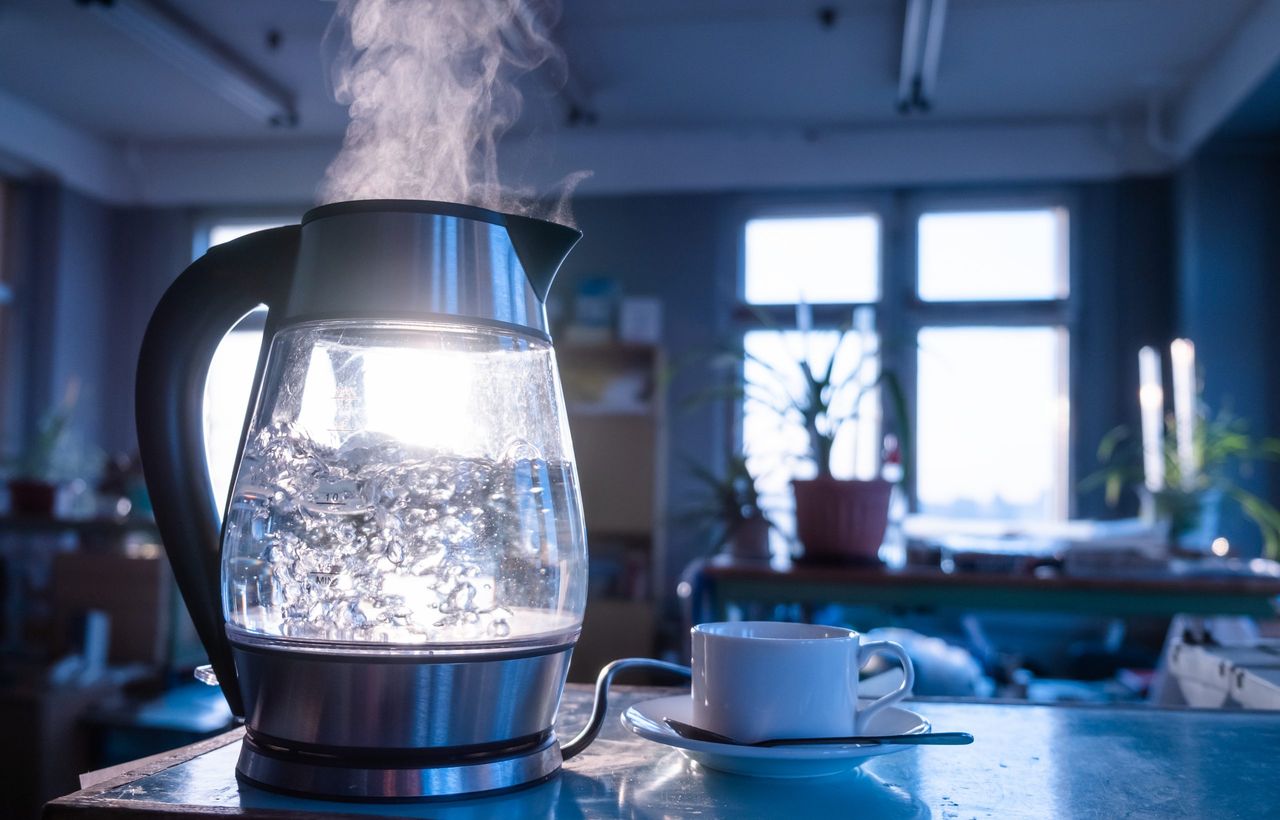 A transparent kettle of water boils against the background of the sunset shining through the window. The concept of coffee break and end of the working day.