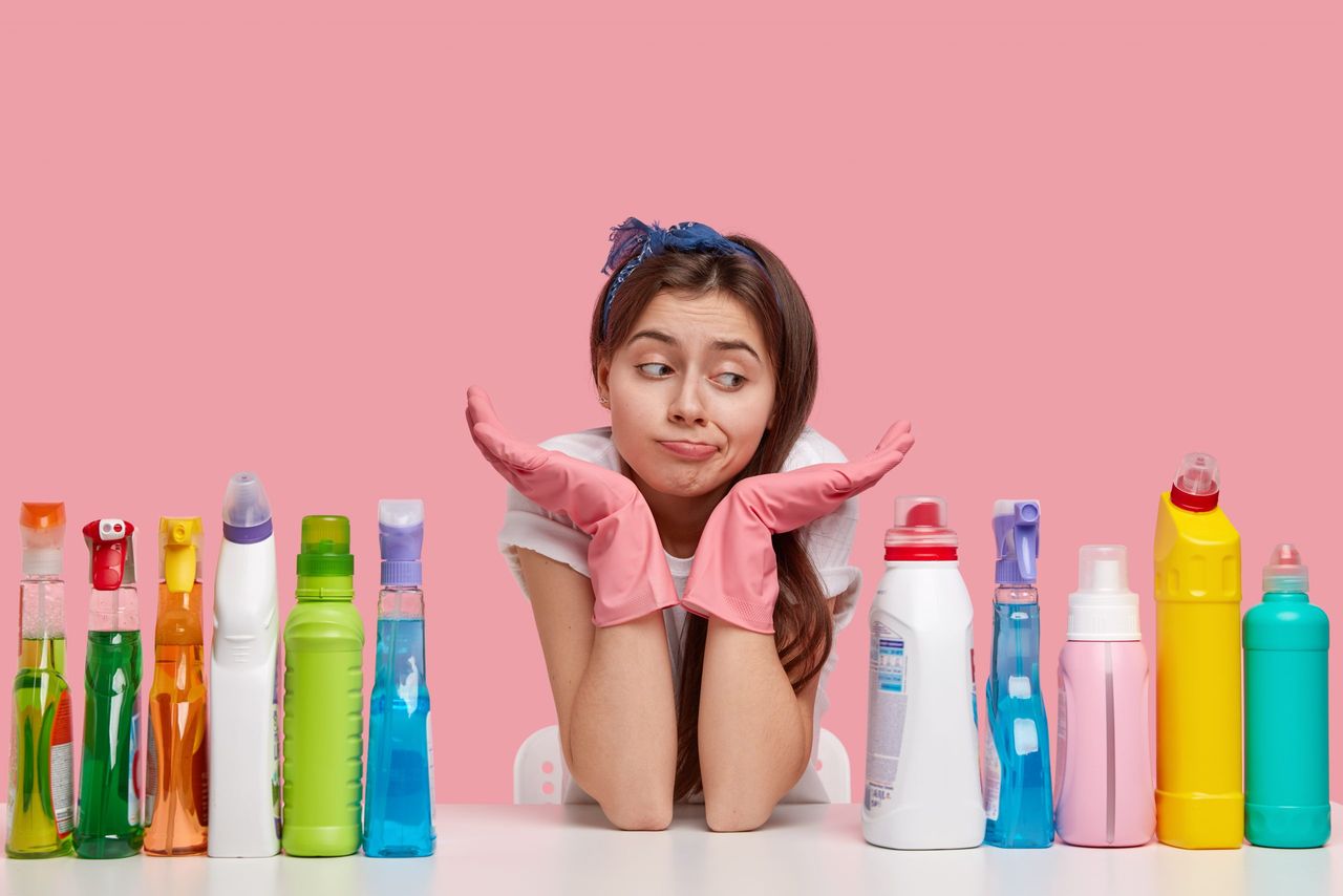 Clueless Caucasian dark haired young woman wears headband, spreads palms, feels apathy towards cleaning house, poses against pink background, feels hesitation. Upset janitor with detergents.
