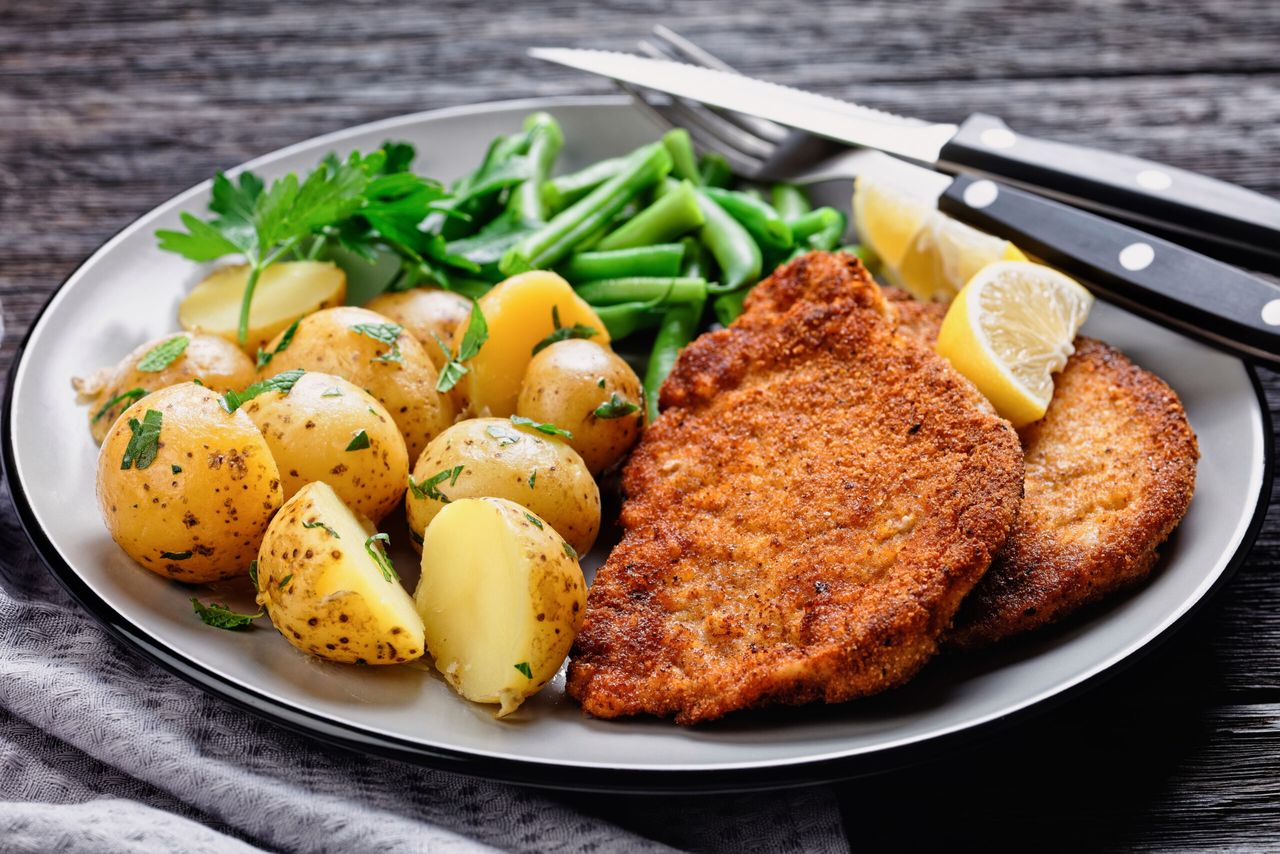 German pork schnitzel with young potatoes, green beans with cutlery served on a plate with lemon wedges, and mayonnaise based sauce on a dark wooden background, top view, close-up