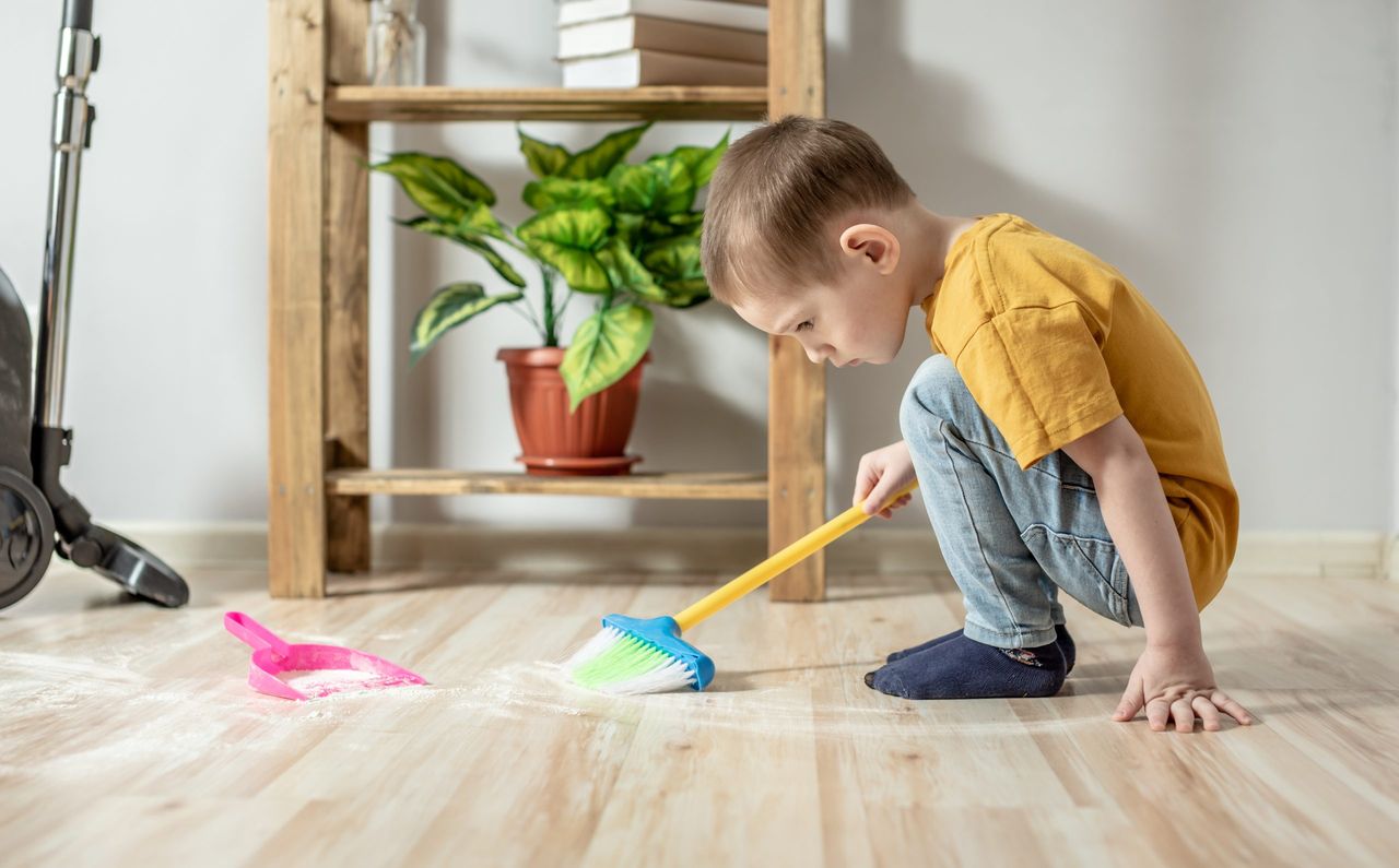 A cute little boy is sweeping the trash from the floor with a broom to a dustpan. Child helps the parents in cleaning the house