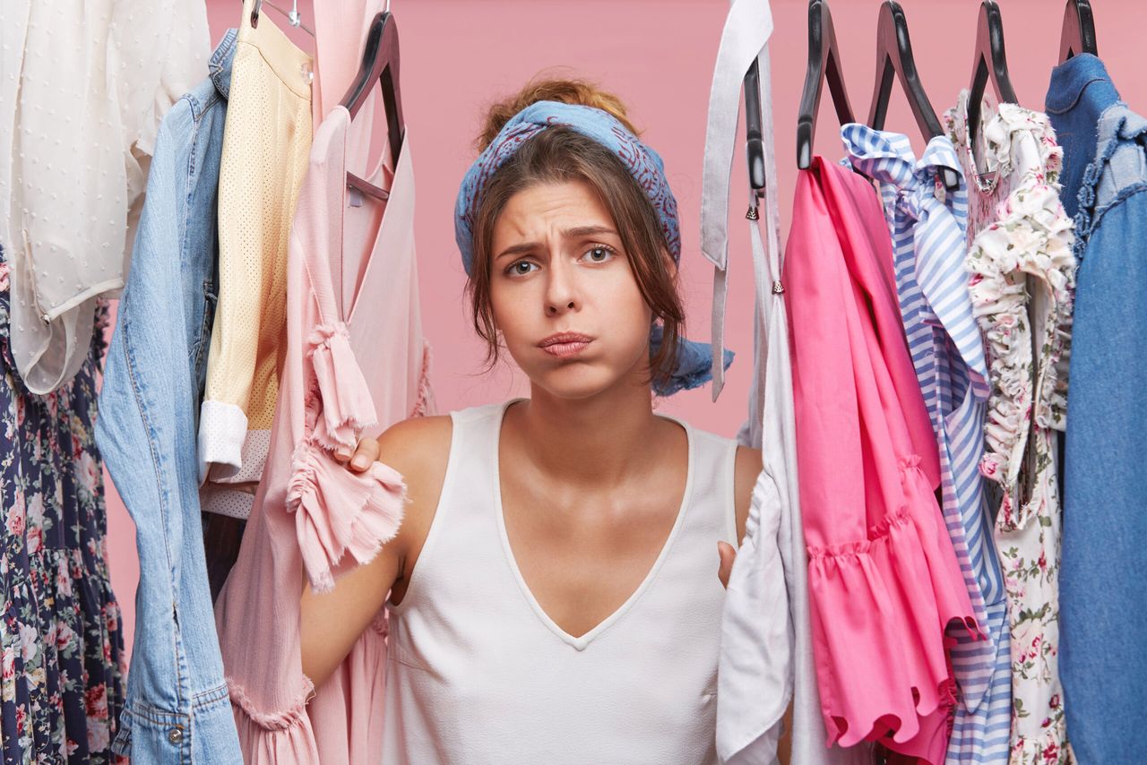 Depressed female standing near wardrobe rack full of clothes, having difficult choice not knowing what to put on. Nothing to wear concept. Choosing clothes for special occasion. Searching for clothing