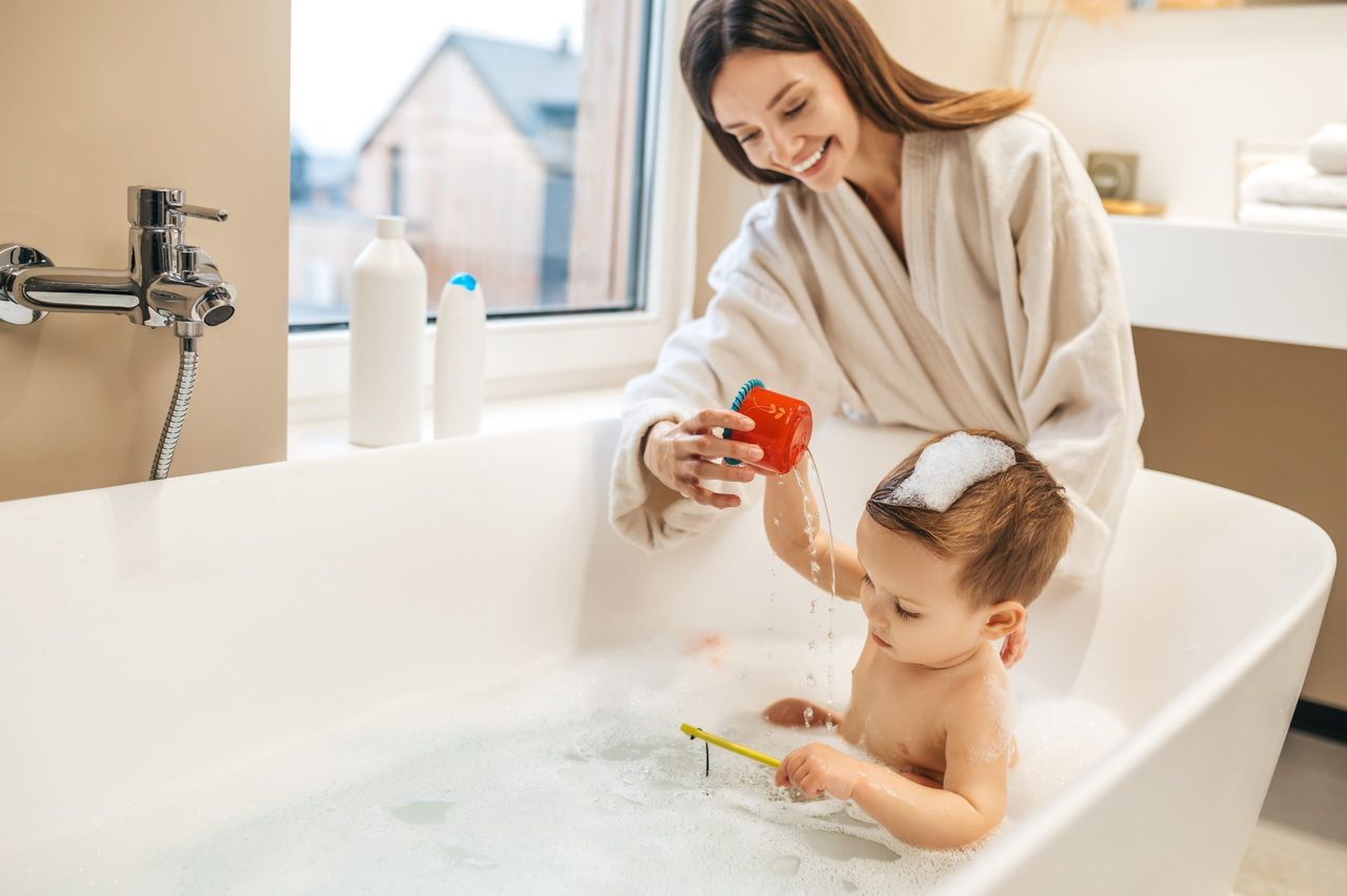 Merry beautiful young female parent bending over her cute baby boy seated in the bath