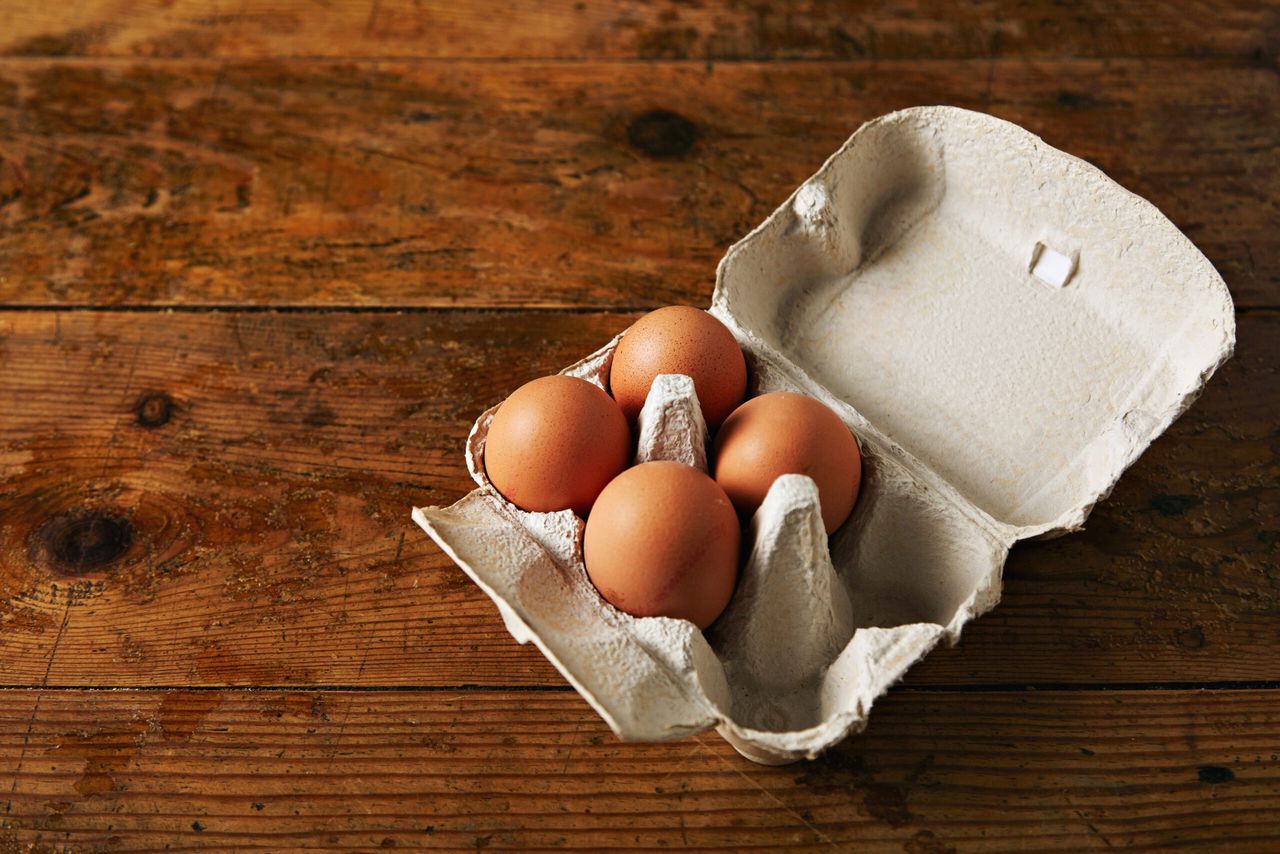 Open egg carton for six eggs containing four brown eggs on a rough rustic brown wooden table